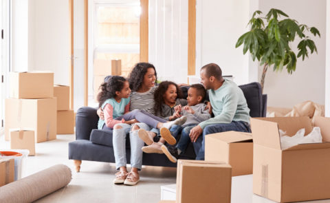 A mother and father and their three children sit together on a couch, surrounded by moving boxes in their living room