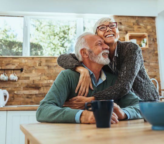 An elderly woman hugging an elderly man in a kitchen