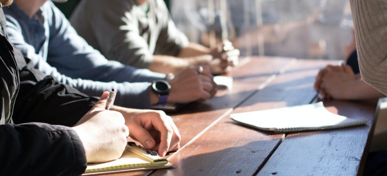 Pairs of hands resting on a table and one pair of hands writing notes on a notepad